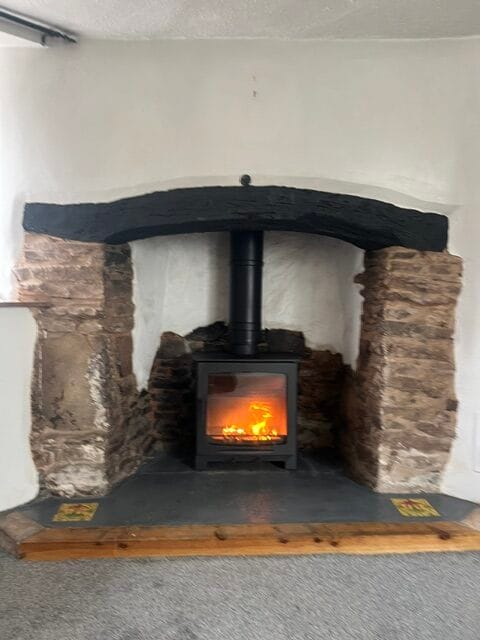 A lit Stirling 5kW wood-burning stove set within a rustic stone fireplace with a black timber lintel and hearth